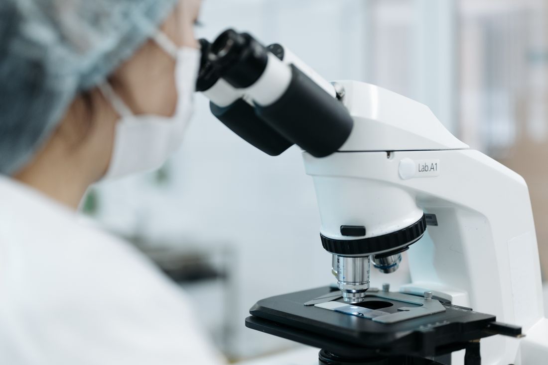 A laboratory technician in a cap and mask looking through a microscope